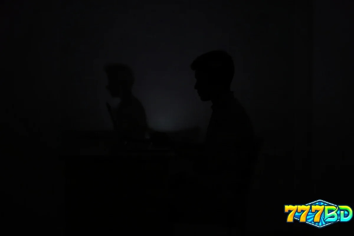 অন্ধকার ঘরে ডেস্কে বসা ব্যক্তি, ল্যাপটপে হাত—অনলাইন নিরাপত্তা যাচাইয়ের দৃশ্য